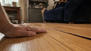A close-up photo of a hand feeling the raised edges of warped hardwood floorboards in a sunlit living room.